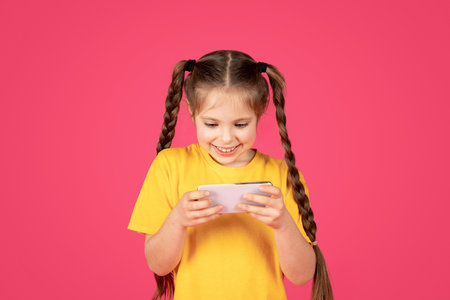 Cheerful preteen girl playing games on smartphone while standing on pink background