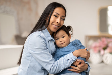 Asian Mom Embracing Infant Daughter Sitting In Cozy Bedroom Interiorの写真素材
