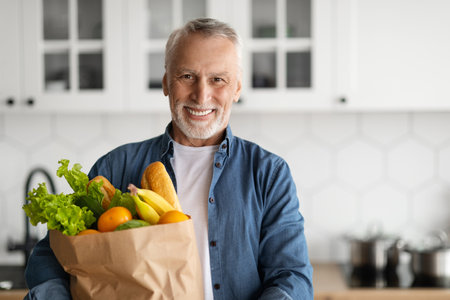 Happy Retirement. Smiling Senior Man Posing With Grocery Bag In Kitchen Interiorの写真素材