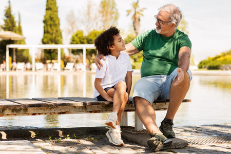 Cheerful senior man with beard and black little boy sitting on bench, relaxing in park, talkの写真素材