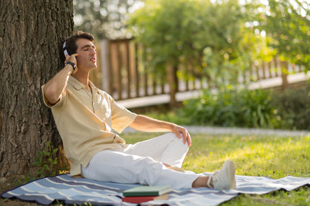 Relaxed young man sitting under tree, listening to musicの写真素材