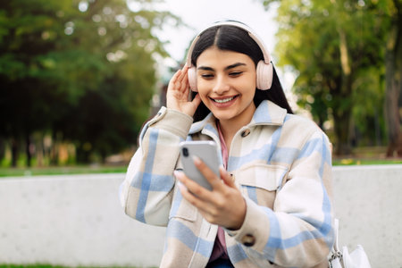 Happy hispanic lady using smartphone and listening music in headphones, texting, sitting outdoors in parkの写真素材