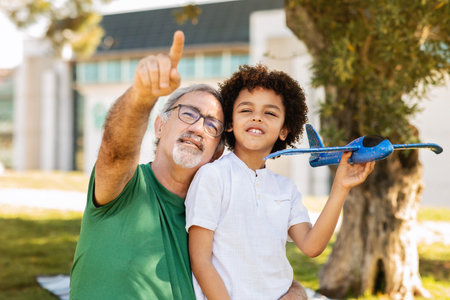 Positive senior man with beard and black little boy hug, have fun in park, flying toy airplane, outdoorの写真素材