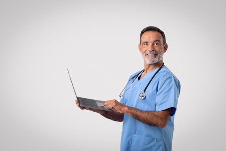 Happy caucasian mature man doctor in blue uniform typing on computer, isolated on gray backgroundの写真素材