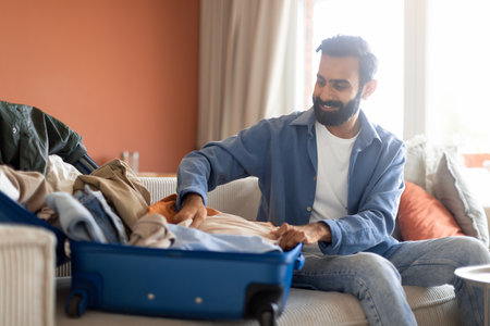 Smiling Indian Guy Packing Travel Suitcase Sitting At Homeの写真素材
