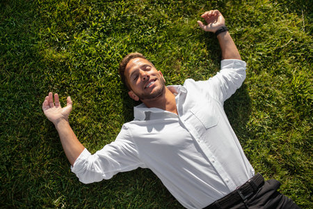 Happy relaxed young businessman in shirt lying on green grass on lawn and resting in park with...