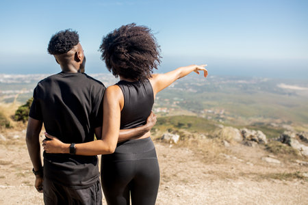 African american lady hugging man enjoying workout break, pointing finger aside, standing on rocks at ocean, outdoorsの写真素材