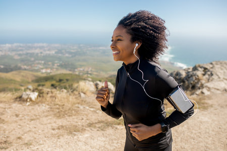 Excited black lady in sports wear, headphones with smartphone running on rocks near ocean alone in...