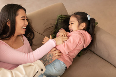 Cheerful Chinese Woman Tickling Little Daughter And Laughing At Homeの写真素材