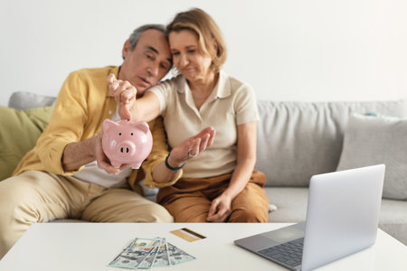 Sad senior spouses counting money from piggybank, sitting on sofa with laptop at homeの写真素材