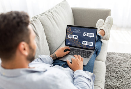 Unrecognizable man sitting on couch, using laptop with Online Assistantの写真素材