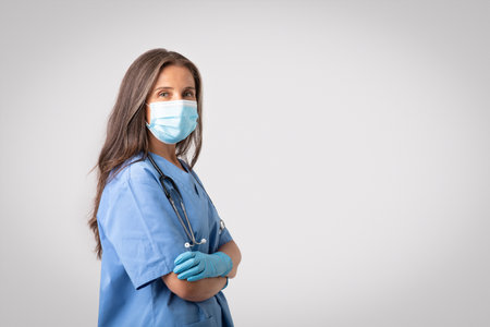 Professional female doctor in protective face mask, posing with folded arms looking at camera over light grey backgroundの写真素材