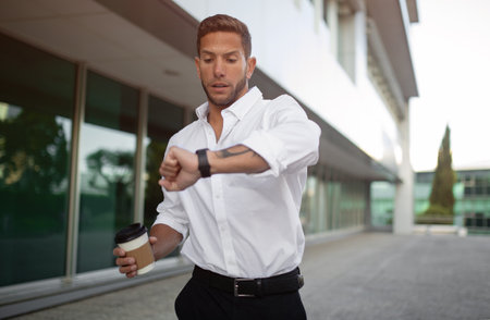 Worried european businessman looking at watch and holding takeaway coffee on the way to office being late for meetingの写真素材