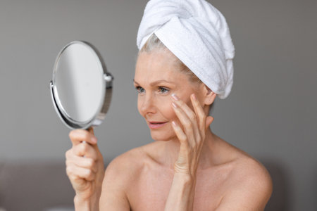 Eye wrinkles treatment. Beautiful mature lady using moisturizing eye cream looking at mirror enjoying face care routineの写真素材