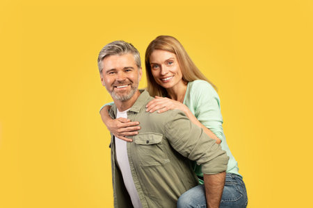 Excited european middle aged couple fooling together, happy woman piggybacking her husband, yellow backgroundの写真素材
