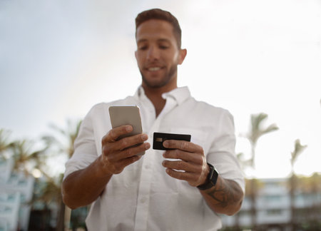 Young businessman with smartphone and credit card in hands standing outdoors, making online shopping or money transfersの写真素材