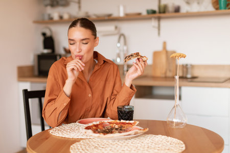 Satisfied woman licking her fingers after tasting delicious pizza slice, enjoying homemade meal in kitchen, free spaceの写真素材