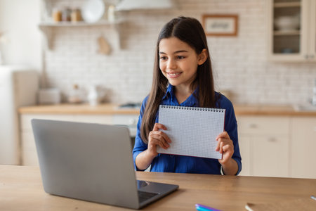 Smiling School Girl Showing Her Copybook To Computer At Home