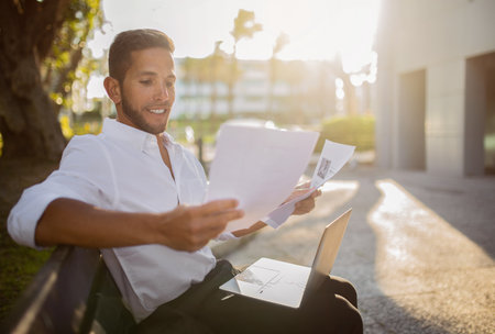 Happy european man entrepreneur working on laptop and reading documents, doing paperwork, sitting on...
