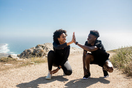 Excited young black couple tying shoelaces, giving high five, enjoy workout together in morning on coast landscapeの写真素材