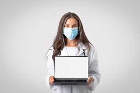 Professional senior doctor woman in white coat, wearing protective mask showing laptop with blank white screen, mockupの写真素材