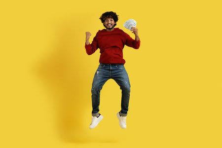 Happy rich young indian guy holding money cash, jumping up