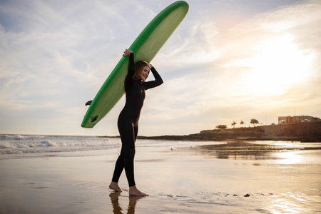 Portrait of young female surfer walking with surfboard at beach on sunset