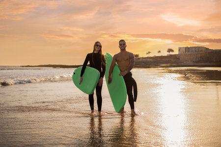 Young Surfers Couple With Surfboards Walking On The Beach At Sunset Time
