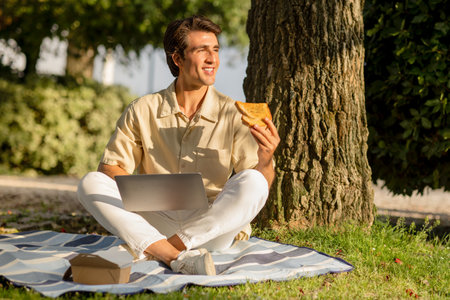 Happy man have picnic at park, using laptop, eating sandwichの写真素材