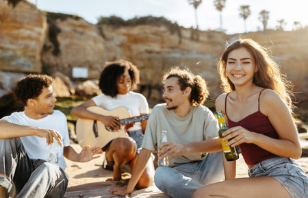 Cheerful millennial african american, asian, european people students with bottles playing guitar, enjoy picnicの写真素材