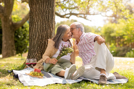 Smiling old european husband gives bouquet of flowers to wife, kiss, enjoy picnic together in parkの写真素材