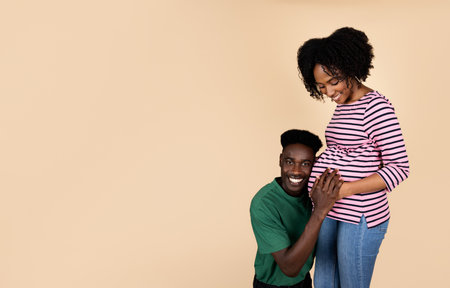 Cheerful young african american husband listening with ear big belly of pregnant wifeの写真素材