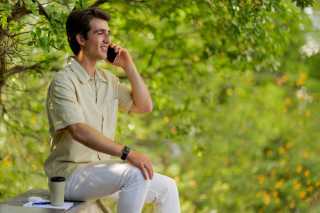 Happy millennial man sitting at park, talking on phoneの写真素材