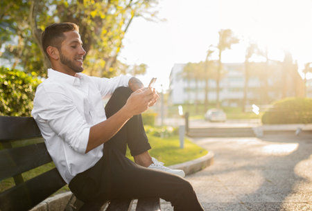 Cheerful young businessman in formal outfit using cellphone and smiling, sitting on bench in park, free spaceの写真素材