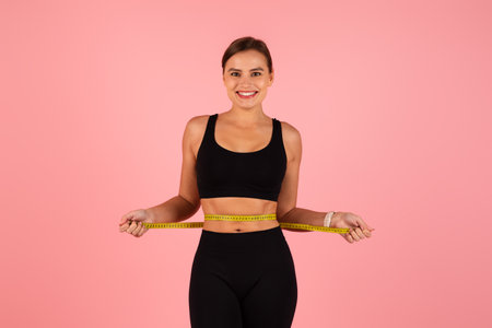 Slimming and dieting. Smiling woman measuring her waist over pink studio backgroundの写真素材