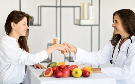 Happy adult european woman doctor nutritionist in white coat shaking hands with young lady patientの写真素材