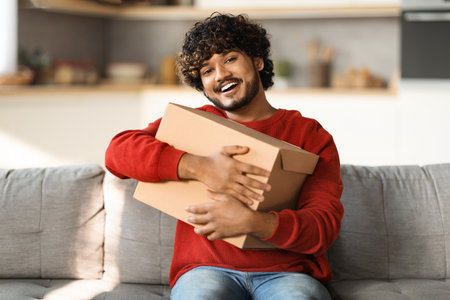 Joyful Young Indian Man Embracing Big Cardboard Box At Homeの写真素材