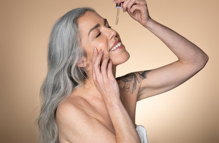 Beautiful smiling senior woman applying facial serum, using anti-aging treatment product, beige studio backgroundの写真素材
