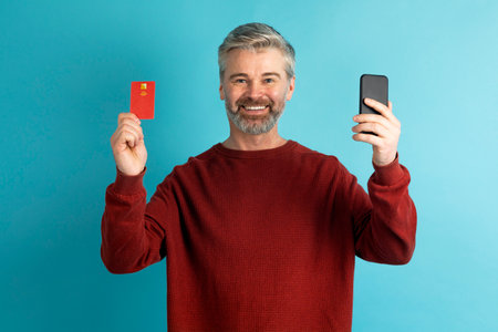 Happy middle aged man showing phone and bank cardの写真素材