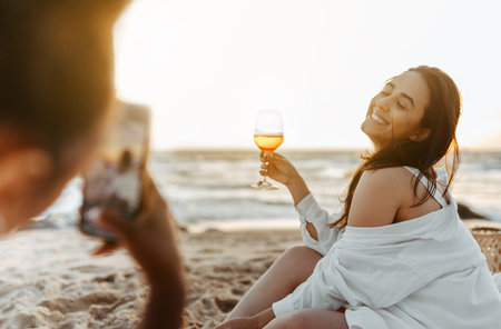 Woman taking a photo of her happy girlfriend at the beach, having picnic and drinking wine at sunset on coaslineの写真素材