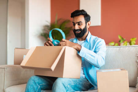 Cheerful middle eastern guy checking delivery package holding headphones indoorの写真素材