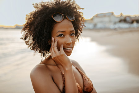 Portrait of young black lady with bushy curly hair in swimwear walking at beach along ocean and posing at cameraの写真素材