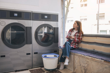Woman Doing Laundry Using Cell Phone Sitting In Laundrette