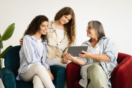 Smiling european adult, senior women and teenager girl sit on sofa, look at tablet in living roomの写真素材