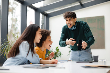 Friends, look. Excited european guy showing cellphone with blank screen to female classmates while...