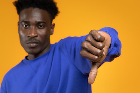 Sad disappointed young black man showing thumb down, yellow background
