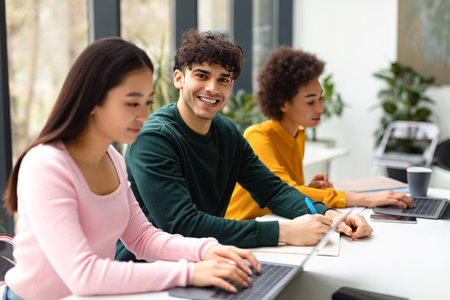 Group of diverse international students sitting at desk in coworking space, focus on smiling guy writing in copybookの写真素材