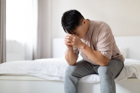 Unhappy middle aged asian man sitting on bed at homeの写真素材