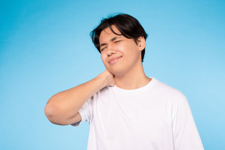 Asian student guy touching neck feeling pain on blue wallの写真素材