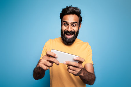 Gadget addiction concept. Happy middle aged indian man playing mobile games on cellphone isolated over blue backgroundの写真素材